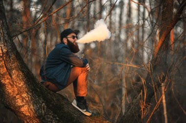 Vape. Güneş gözlüğü ve bir kap içinde büyük sakallı acımasız bir genç adam günbatımında ormanda elektronik sigara sigara içiyor. Yaşam tarzı.