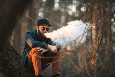 Vape. Güneş gözlüğü ve bir kap içinde büyük sakallı acımasız bir genç adam günbatımında ormanda elektronik sigara sigara içiyor. Yaşam tarzı.