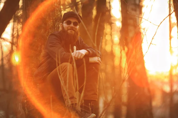 Vape. Un joven brutal con una barba enorme en gafas de sol y una gorra y con cigarrillo electrónico en el bosque al atardecer. Estilo de vida . — Foto de Stock