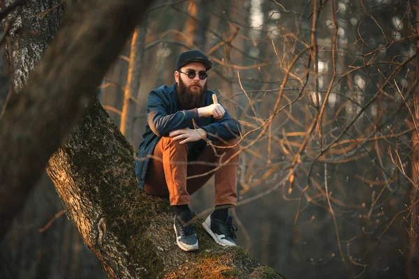 Vape. Een brutale jonge man met een grote baard in zonnebril en een pet en elektronische sigaret in de bossen bij zonsondergang. Levensstijl. — Stockfoto