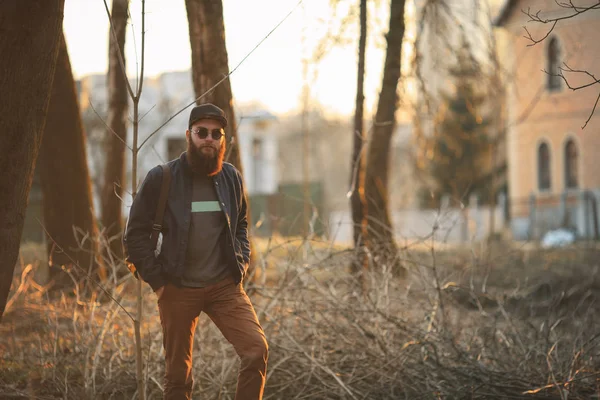A brutal young man with a huge beard in sunglasses and a cap in the woods at sunset. Lifestyle. — Stock Photo, Image