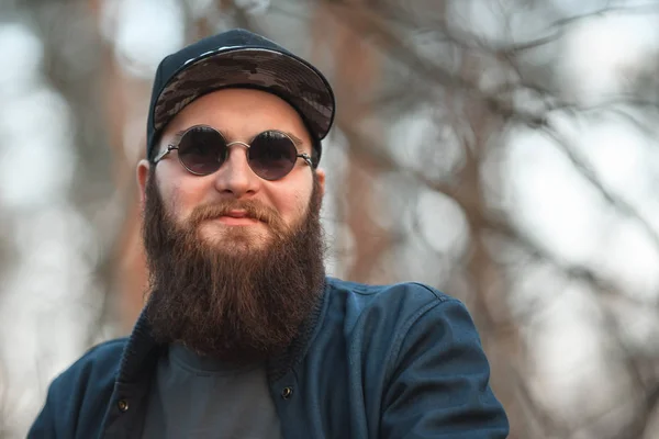 A brutal young man with a huge beard in sunglasses and a cap in the woods at sunset. Lifestyle. — Stock Photo, Image