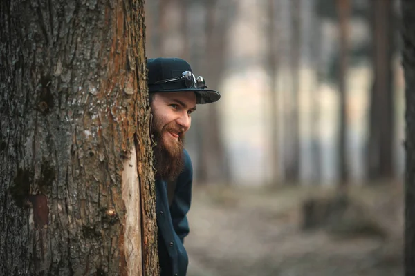 Brutální mladý muž s obrovskou vousy v sluneční brýle a čepici v lese při západu slunce. Životní styl. — Stock fotografie