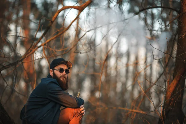 Vape. Een brutale jonge man met een grote baard in zonnebrillen en een cap is het roken van een elektronische sigaret in de bossen bij zonsondergang. Levensstijl. — Stockfoto