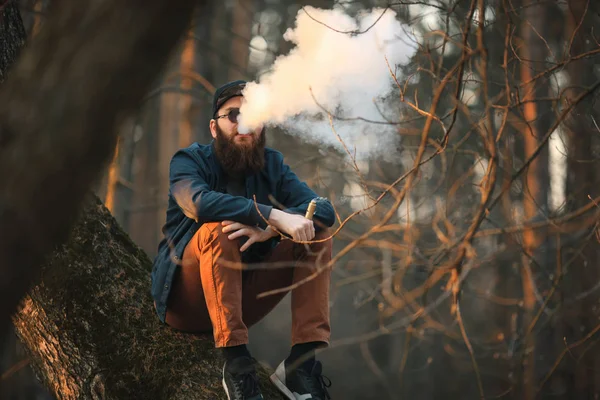 Vape. Un joven brutal con una barba enorme en gafas de sol y una gorra está fumando un cigarrillo electrónico en el bosque al atardecer. Estilo de vida . — Foto de Stock