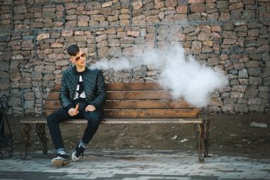 Vape. Genç yakışıklı adam bankta oturur ve elektronik sigara buhar esiyor. Yaşam tarzı.