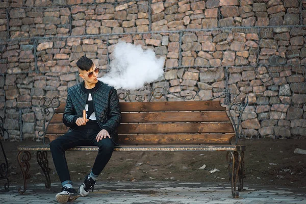 Vape. Genç yakışıklı adam bankta oturur ve elektronik sigara buhar esiyor. Yaşam tarzı. — Stok fotoğraf