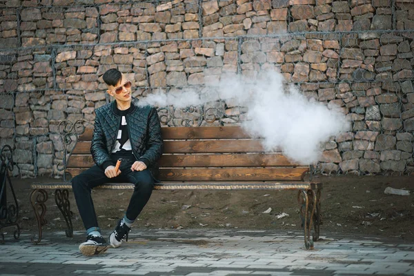 Vape. A young handsome guy sits on the bench and blows steam from an electronic cigarette. Lifestyle. — Stock Photo, Image