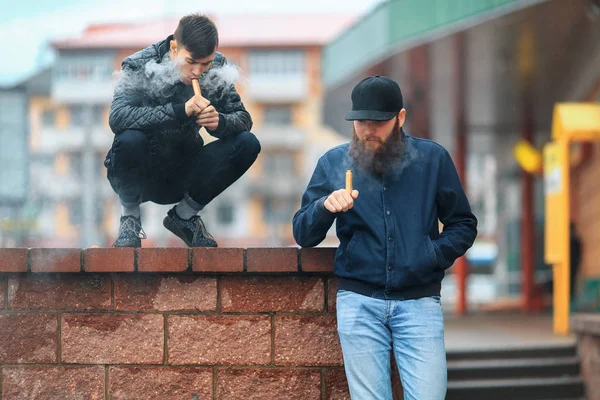 Vape. Two young people stand and smoke electronic cigarettes in the street on a cloudy day.