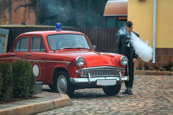 Vape. ein junger gutaussehender Kerl steht neben einem alten Feuerwehrauto und lässt Dampf aus einer elektronischen Zigarette ab. — Stockfoto