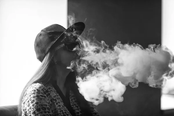 Vape. Jeune belle fille blanche dans des lunettes de soleil et dans une casquette admire les bouffées de vapeur de la cigarette électronique . — Photo