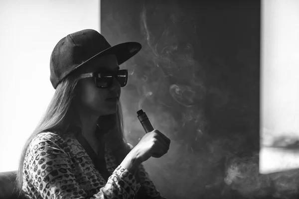 Vape. Menina branca bonito jovem em óculos de sol e em um boné está admitindo sopros de vapor do cigarro eletrônico . — Fotografia de Stock