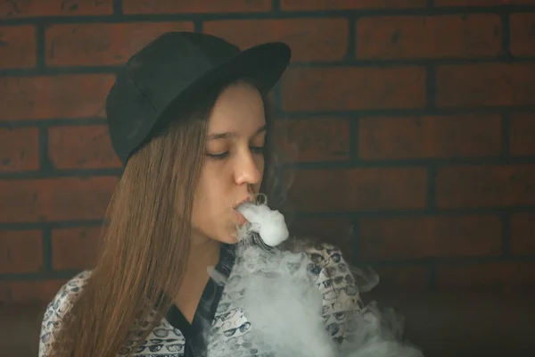 Vape. Young handsome white girl in a cap is admiting puffs of steam from the electronic cigarette.