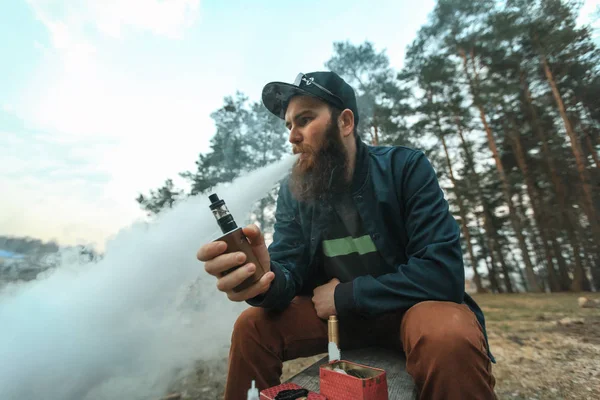Vape. Junger Mann mit langem Bart und Schirmmütze raucht im Wald eine elektronische Zigarette. — Stockfoto