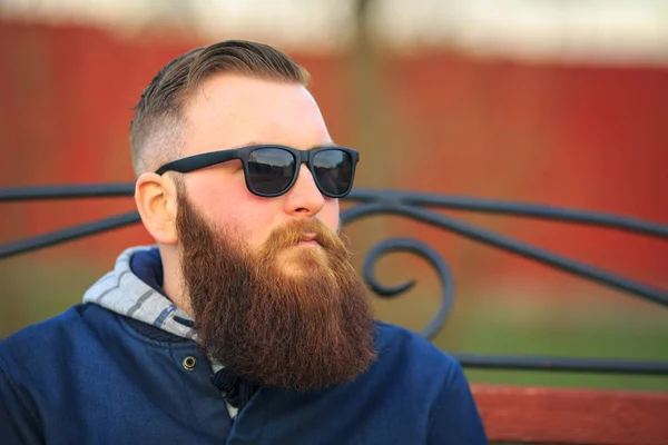 Vape. Young brutal man with large beard and fashionable haircut in sunglasses smokes an electronic cigarette on the red bench in the city park. — Stock Photo, Image