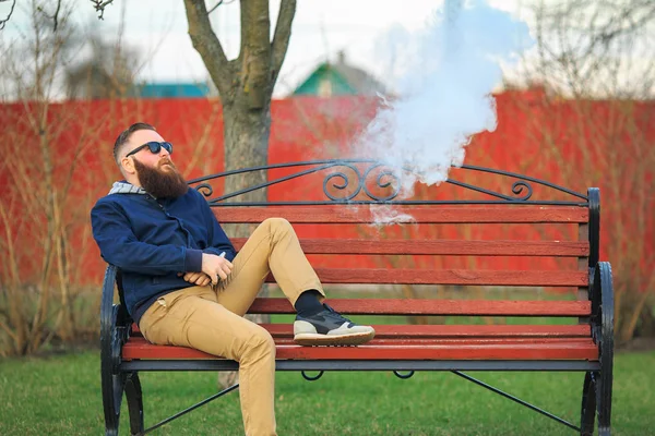Vape. Joven hombre brutal con barba grande y corte de pelo de moda en gafas de sol fuma un cigarrillo electrónico en el banco rojo en el parque de la ciudad . — Foto de Stock