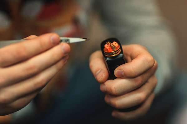Vape after-sales service of the e-cigarette. Male hands regulate electronic cigarette coils with pincette. Glowing spiral.