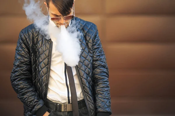 Vape man. Portrait of a handsome young white guy with modern haircut in aviator sunglasses vaping an electronic cigarette opposite the futuristic urban background. — Stock Photo, Image