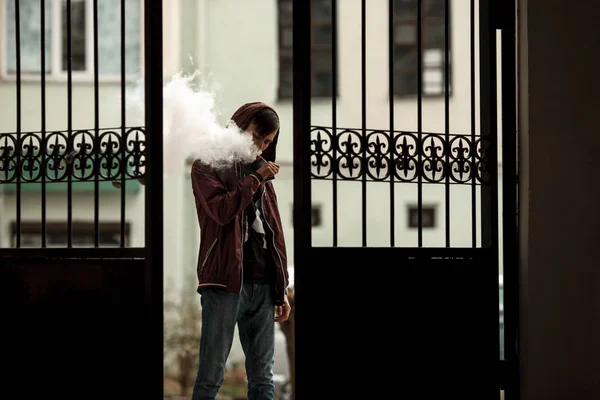 Vape man. Photo of a handsome young white guy in the hood vaping and letting off steam from an electronic cigarette near a vintage metal gate. — Stock Photo, Image