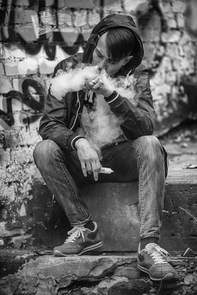 Homem-macaco. Foto de um belo jovem branco no capô vaporizando e soltando vapor de um cigarro eletrônico em uma velha escada de concreto quebrado. Foto em preto e branco . — Fotografia de Stock