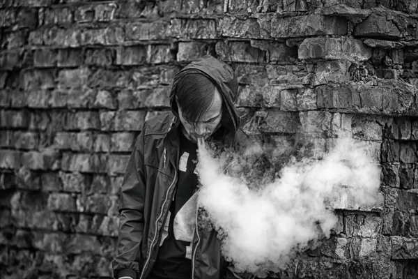 Homem-macaco. Foto de um belo jovem branco no capô vaporizando e soltando vapor de um cigarro eletrônico perto da velha parede de tijolos destruída. Foto em preto e branco . — Fotografia de Stock