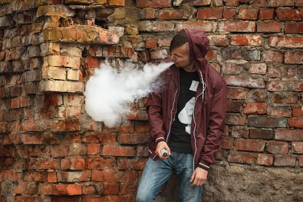 Vape man. Photo of a handsome young white guy in the hood vaping and letting off steam from an electronic cigarette near old destroyed red brick wall. — Stock Photo, Image