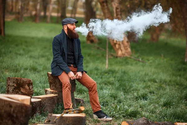 Hombre Vape. Foto de un joven barbudo brutal descansando después de cortar leña y vapear un cigarrillo electrónico en el pueblo . — Foto de Stock