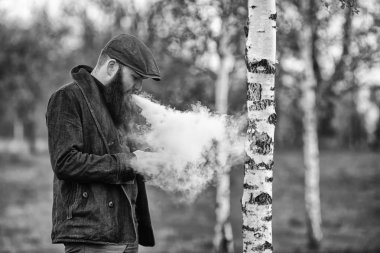 Vape gadget adam. Genç acımasız beyaz bir adam ve onun smartphone köy huş Grove'da bakarak bir vintage cap vaping elektronik sigara ve büyük sakal ile açık portresi. Siyah ve beyaz.