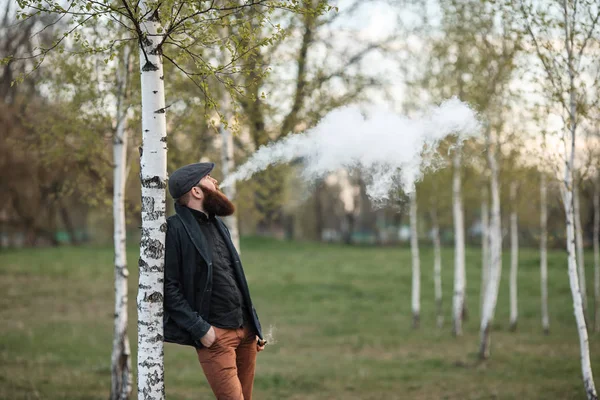 Hombre Vape. Retrato al aire libre de un joven blanco brutal con barba grande y con una gorra vintage que deja soplos de vapor de un cigarrillo electrónico en el abedul en el pueblo . — Foto de Stock