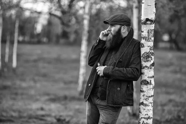 Vape gadget man. Outdoor Portret van een jonge brute blanke man met grote baard en in een vintage cap vaping een elektronische sigaret en praten over de smartphone in de berk grove in het dorp. Zwart-wit. — Stockfoto