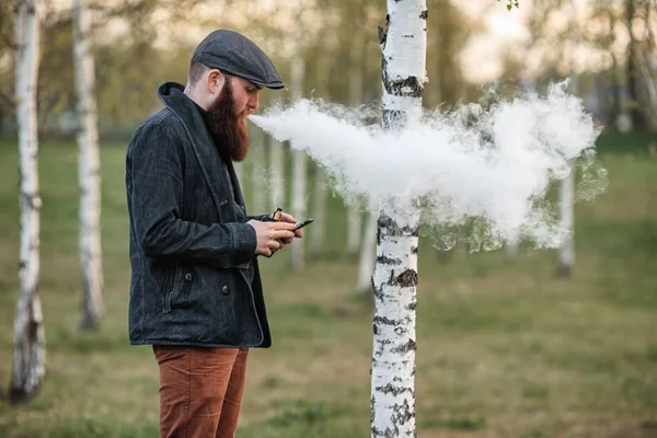 Vape gadget adam. Açık bir genç acımasız beyaz adam büyük sakallı ve vintage cap vaping elektronik sigara ve onun smartphone köy huş Grove'da bakarak portresi. — Stok fotoğraf