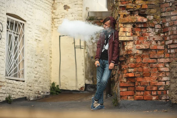 Vape adam. Hood vaping ve eski yıkılan kırmızı tuğla duvar yakınındaki elektronik sigara buhar kapalı icar yakışıklı genç bir beyaz adam fotoğrafı.