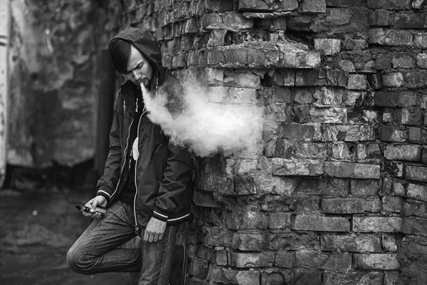 Homem-macaco. Foto de um belo jovem branco no capô vaporizando e soltando vapor de um cigarro eletrônico perto da velha parede de tijolos destruída. Foto em preto e branco . — Fotografia de Stock