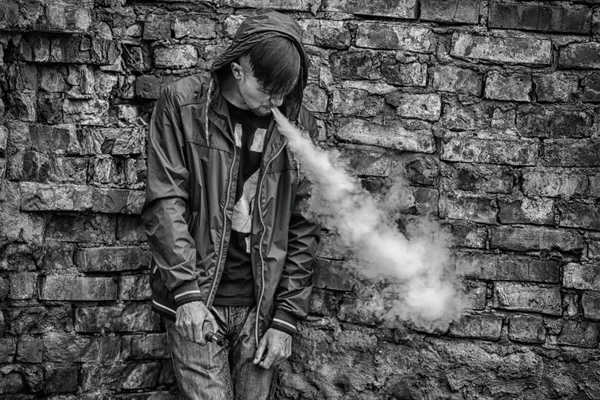 Homem-macaco. Foto de um belo jovem branco no capô vaporizando e soltando vapor de um cigarro eletrônico perto da velha parede de tijolos destruída. Foto em preto e branco . — Fotografia de Stock
