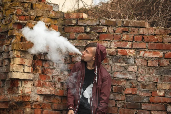 Vape man. Photo of a handsome young white guy in the hood vaping and letting off steam from an electronic cigarette near old destroyed red brick wall. — Stock Photo, Image