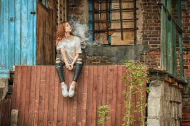 Güzel vape genç. Kırmızı kıvırcık saçlı vaping eski harap köy evinin yakınındaki elektronik sigara ile genç ve güzel bir beyaz kız portresi.