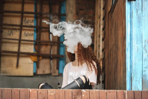 Nastolatek piękny vape. Portret dość młoda dziewczyna biały z rude włosy kręcone vaping elektroniczny papieros w pobliżu stary zniszczony dom. — Zdjęcie stockowe