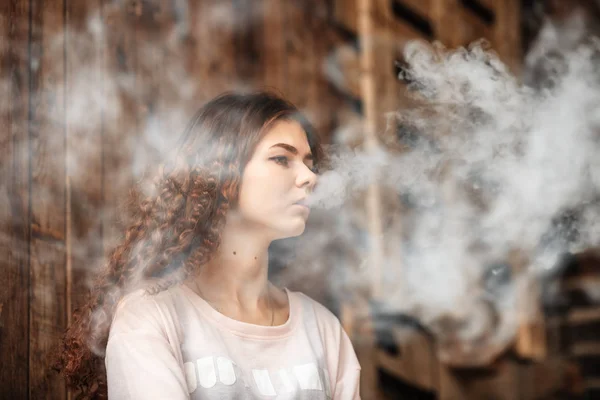 Beautiful vape teenager. Portrait of a pretty young white girl with red curly hair vaping an electronic cigarette near the old ruined village house. — Stock Photo, Image