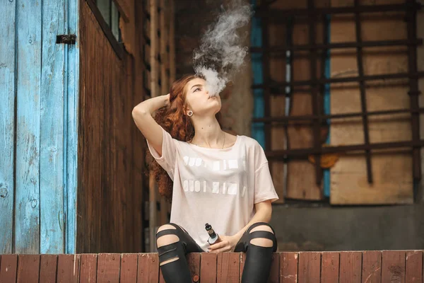 Güzel vape genç. Kırmızı kıvırcık saçlı vaping eski harap köy evinin yakınındaki elektronik sigara ile genç ve güzel bir beyaz kız portresi. — Stok fotoğraf