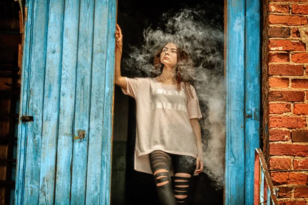 Mooie vape tiener. Portret van een mooie jonge witte meisje met rood krullend haar vaping een elektronische sigaret in de buurt van de oude verwoeste dorpshuis. — Stockfoto