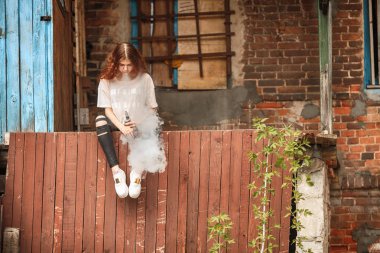 Güzel vape genç. Kırmızı kıvırcık saçlı vaping eski harap köy evinin yakınındaki elektronik sigara ile genç ve güzel bir beyaz kız portresi.