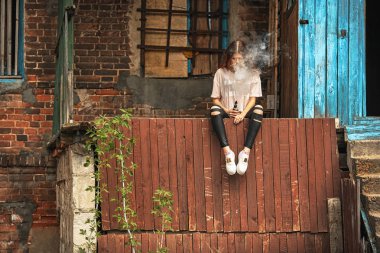 Güzel vape genç. Kırmızı kıvırcık saçlı vaping eski harap köy evinin yakınındaki elektronik sigara ile genç ve güzel bir beyaz kız portresi.