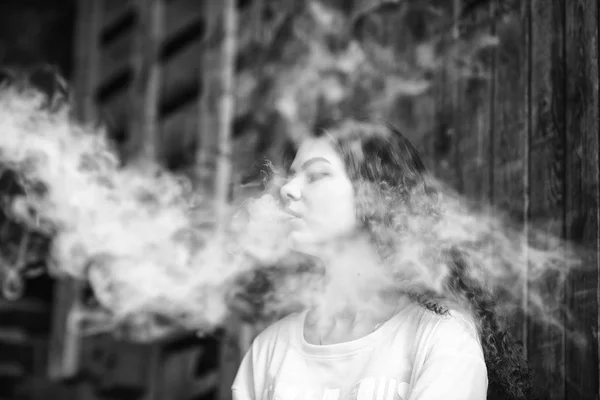 Bela adolescente vape. Retrato de uma menina branca bonita com cabelo encaracolado vaping um cigarro eletrônico perto da antiga casa da aldeia arruinada. Preto e branco . — Fotografia de Stock