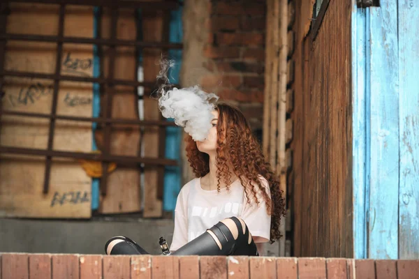 Bela adolescente vape. Retrato de uma jovem menina branca bonita com cabelo encaracolado vermelho vaping um cigarro eletrônico perto da antiga casa da aldeia arruinada . — Fotografia de Stock