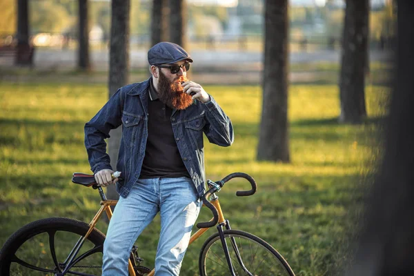 Vape in real life. Young man with gourgeous large beard and in sunglasses and in the cap having a rest and vaping an electronic cigarette near vintage fix bicycle after ride in the city park.