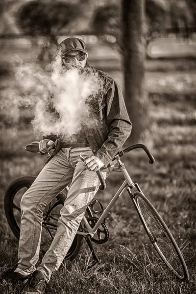 Vape na vida real. Jovem com barba grande e elegante e óculos de sol e na tampa tendo um descanso e vaping um cigarro eletrônico perto de bicicleta correção vintage após passeio no parque da cidade. Tonificado . — Fotografia de Stock