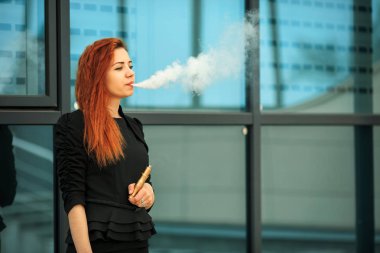 Vape genç. Şehir arka plan karşısında elektronik sigara vaping elbise genç güzel beyaz kız kırmızı kıvırcık saçlı ve siyah modern saç kesimi.