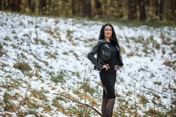 Söt vit flicka med svart hår och svarta kläder poserar mot kanten av skogen med snö under våren. — Stockfoto