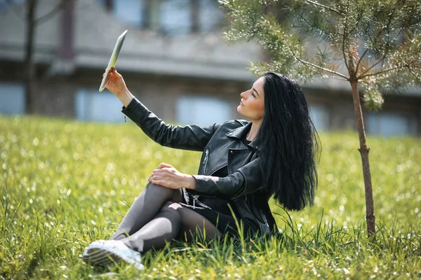 Selfie. Joven chica blanca bonita con el pelo negro y en ropa negra sentado en la hierba verde y navegar en Internet en la tableta blanca . — Foto de Stock