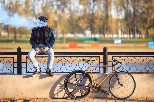 Vape in real life. Young man with gourgeous large beard and in sunglasses and in the cap having a rest and vaping an electronic cigarette near vintage fix bicycle after ride on the waterfront. — Stock Photo, Image